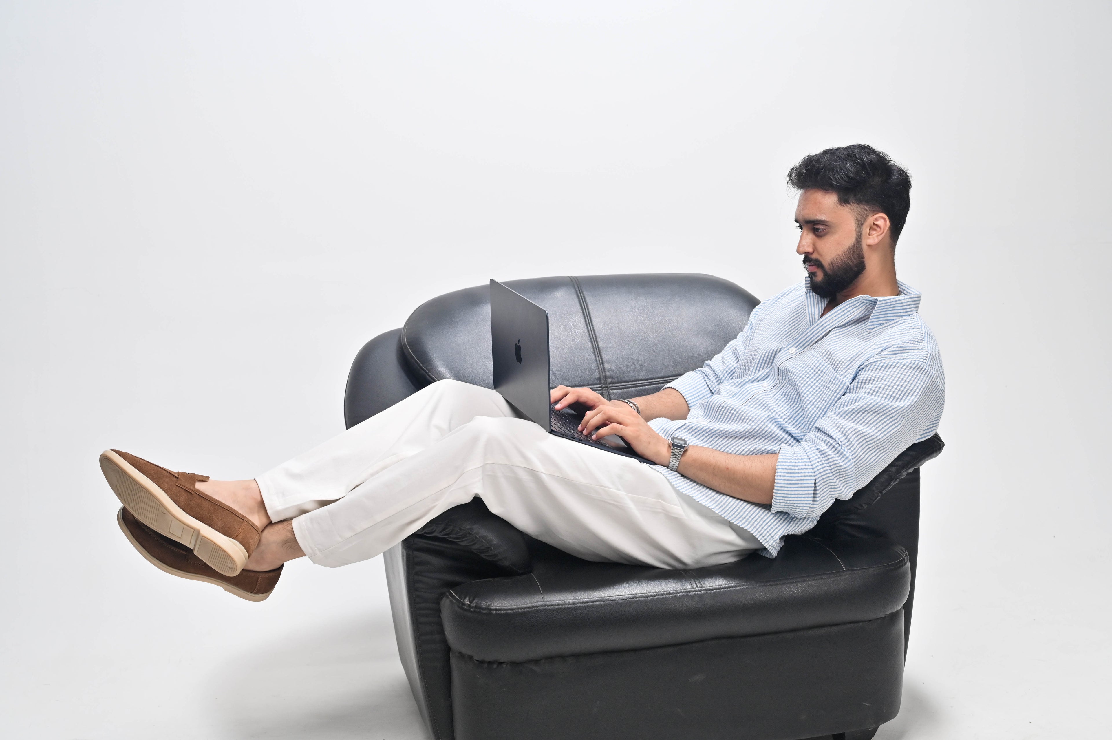 A man with a beard sits in a black leather armchair, typing on a laptop placed on his lap. He is wearing a light blue button-up shirt, off-white trousers, and brown slip-on shoes against a plain white studio background.