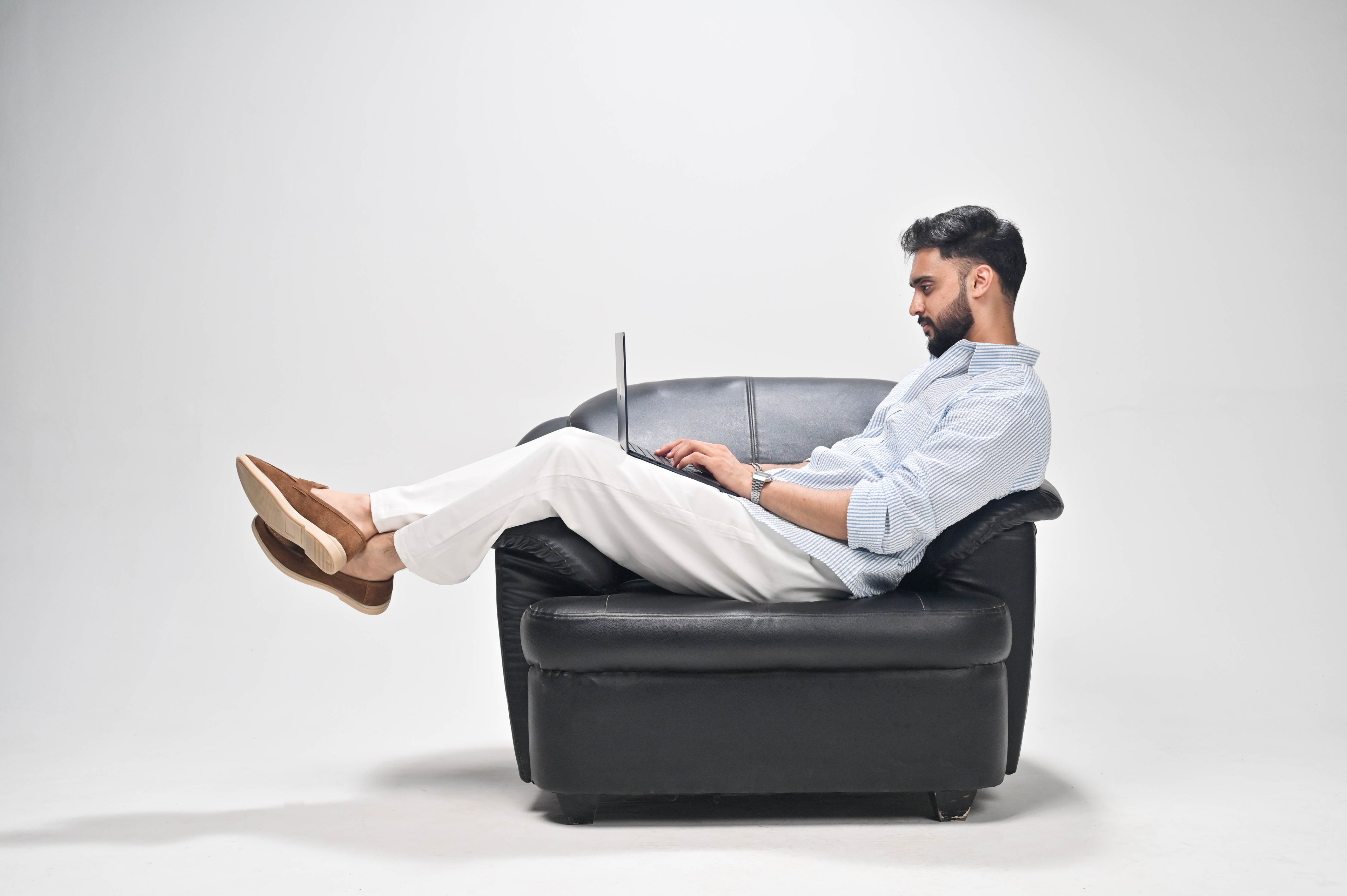 A man with a beard sits in a black leather armchair, using a laptop with his legs stretched out. He is wearing a light blue button-up shirt, white trousers, and brown loafers, set against a plain light gray studio background.