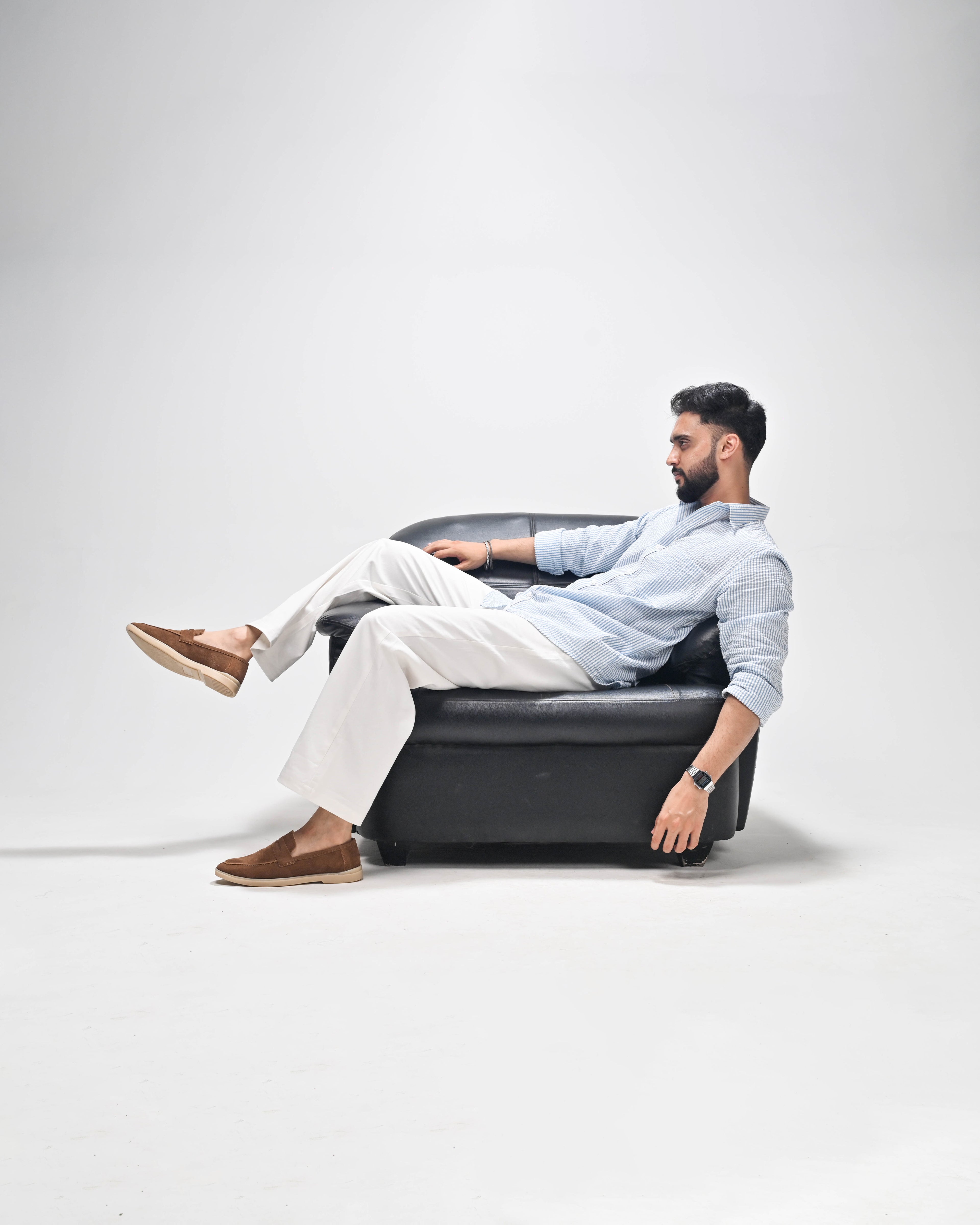 A man wearing a light blue button-down shirt, white trousers, and brown loafers sits relaxed on a black leather armchair against a plain light gray studio background.
