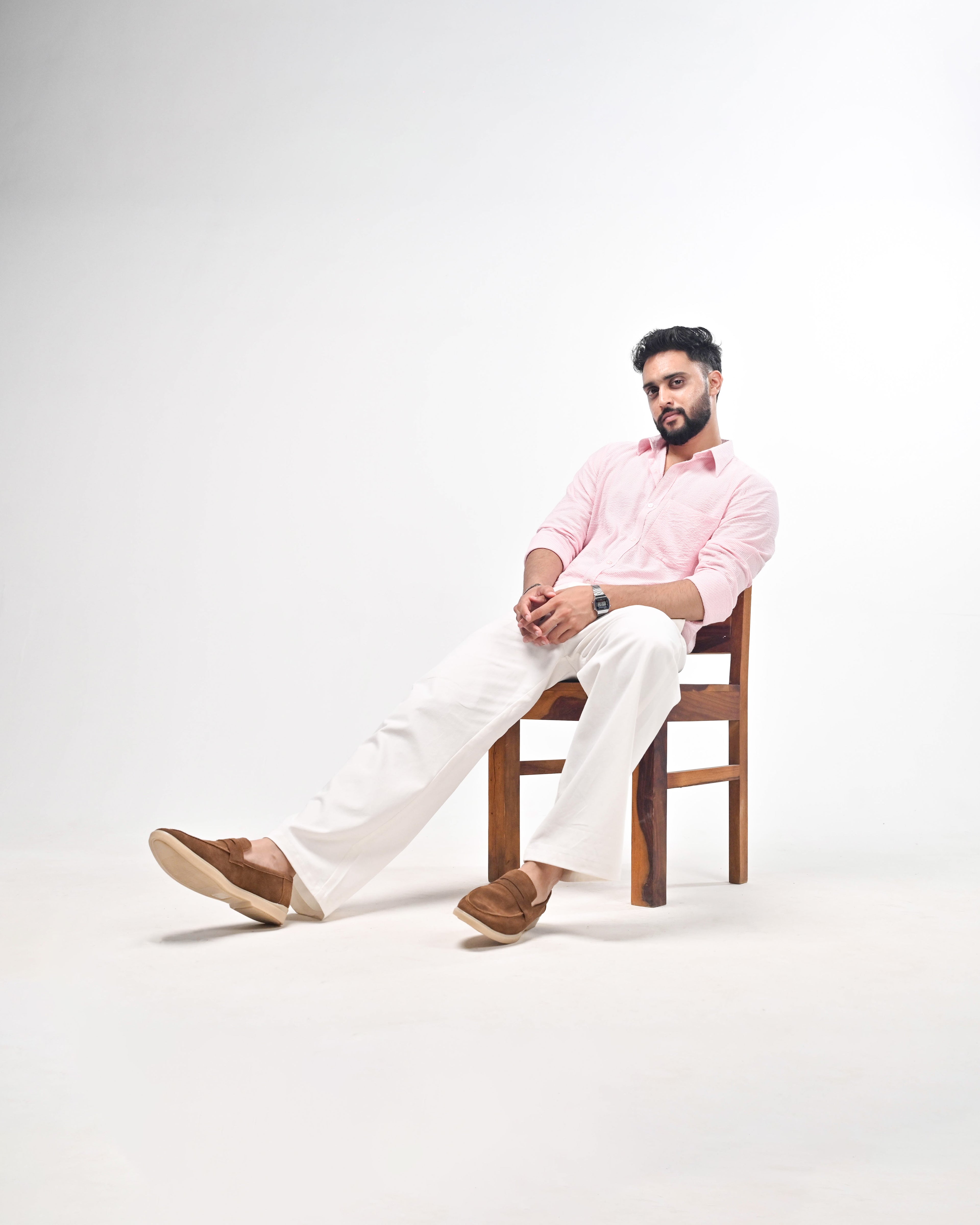 A man wearing a light pink long-sleeve button-up shirt, white trousers, and brown suede loafers sits on a wooden chair against a clean white studio background.